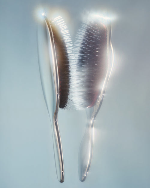 Two translucent hair brushes with dark bristles, shown against a soft light blue-gray background with bright light flares.