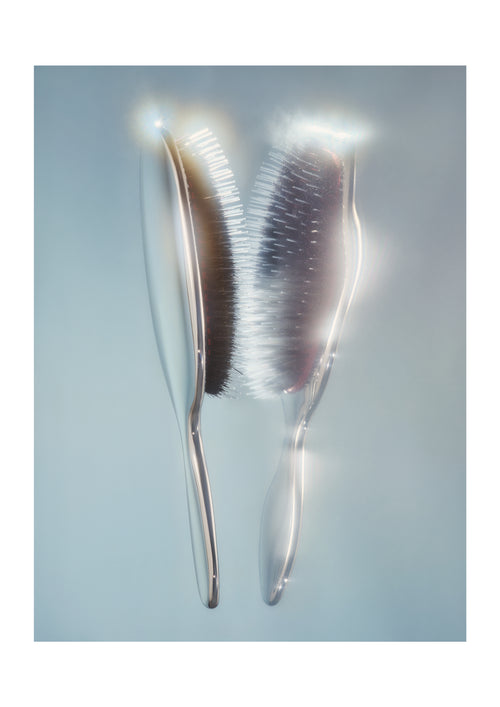 Two translucent hairbrushes with silver handles and dark bristles, shown against a soft light blue background with bright light flares.
