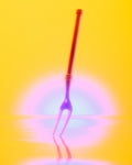 A neon-colored two-pronged fork standing upright on a reflective yellow surface against a glowing yellow background with pink and blue light effects.