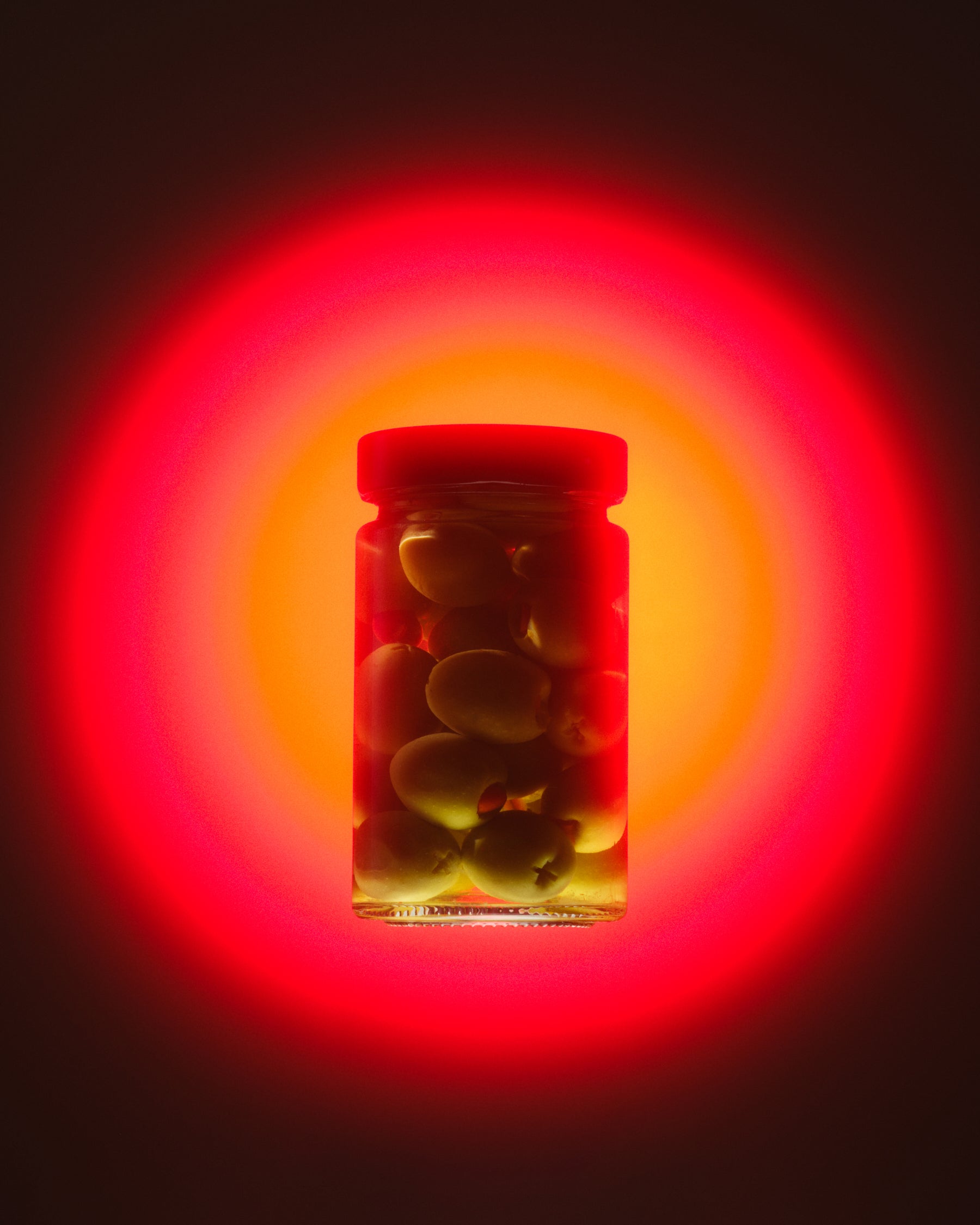 A clear glass jar filled with green olives on the pit, set against a glowing red and orange circular light effect on a dark background.