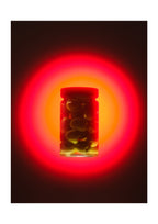 A glass jar filled with dark olives or similar round food items, centered against a black background with a dramatic red and orange glow behind it.