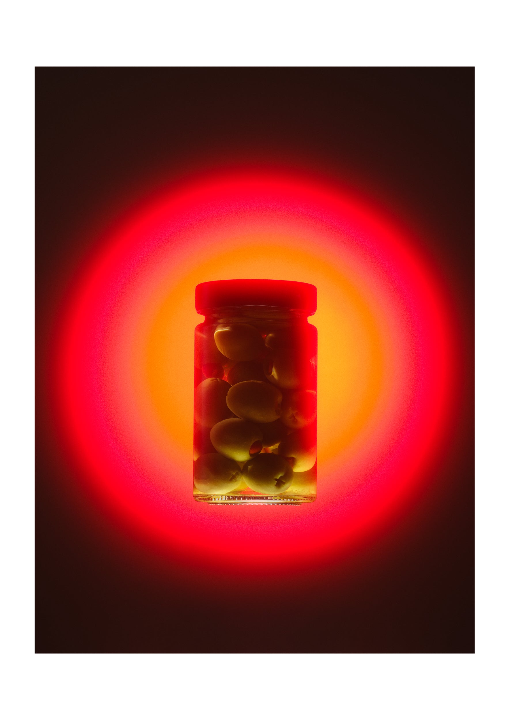 A glass jar filled with dark olives or similar round food items, centered against a black background with a dramatic red and orange glow behind it.