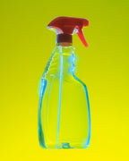 A clear plastic spray bottle with a red trigger sprayer, shown against a bright yellow gradient background.