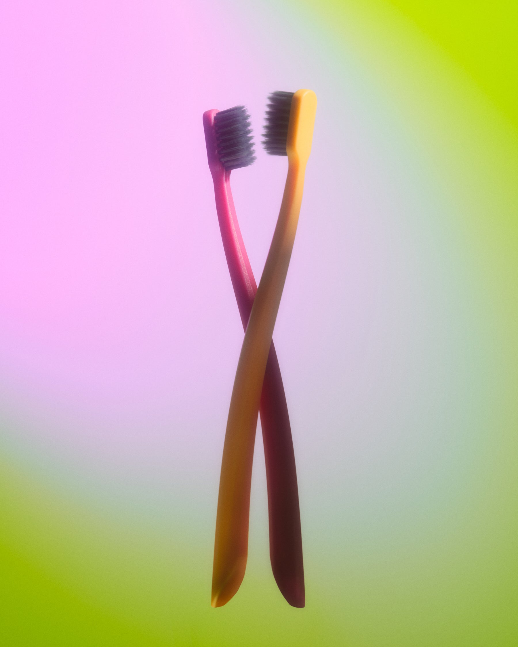 Two crossed toothbrushes with pink and yellow handles, standing upright against a soft pastel gradient background that shifts from pink to green.